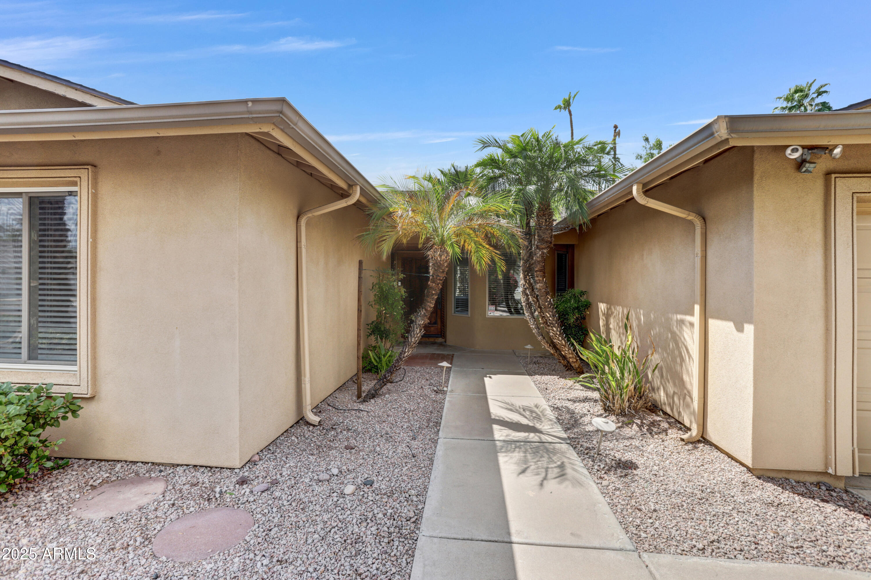 Tan stucco home entrance with a palm-lined walkway through a gravel courtyard.