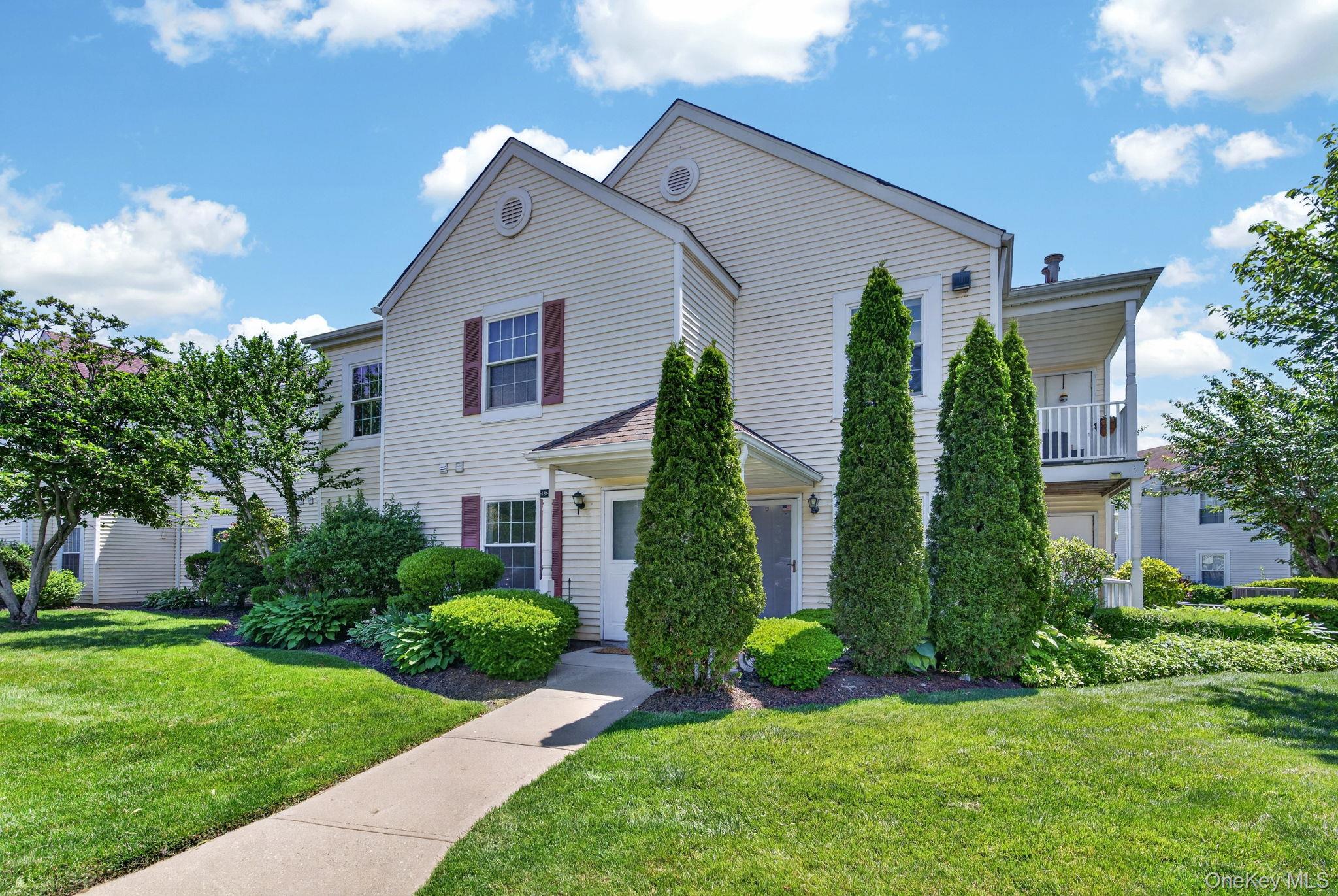 Two-story light-sided house with burgundy shutters, tall evergreen shrubs, and a landscaped front lawn.