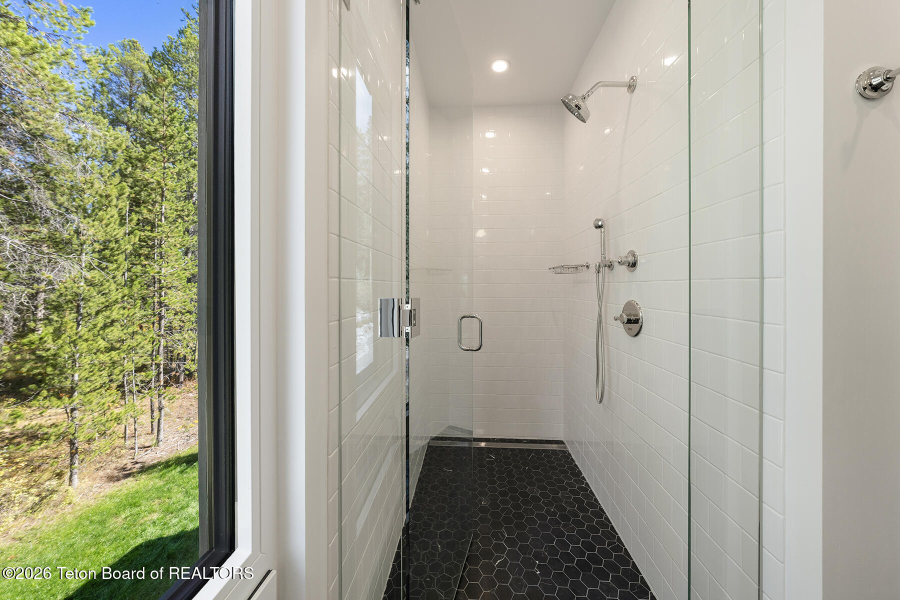 Walk-in shower with white tile, glass enclosure, chrome fixtures, and a window overlooking trees.