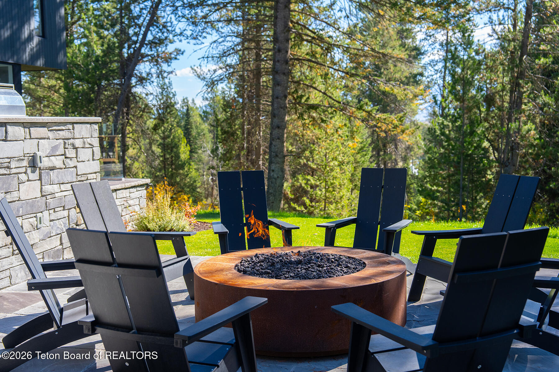 Stone patio with dark chairs arranged around a circular fire pit, backed by a wooded lawn.