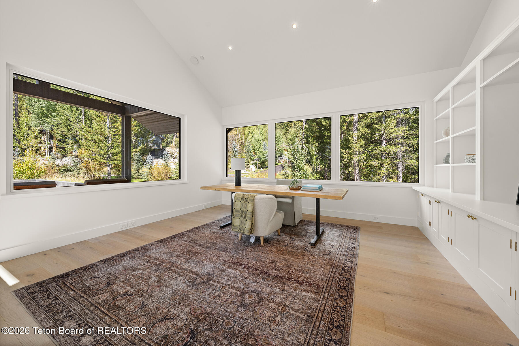 Bright home office with a long wooden desk and large windows overlooking trees.