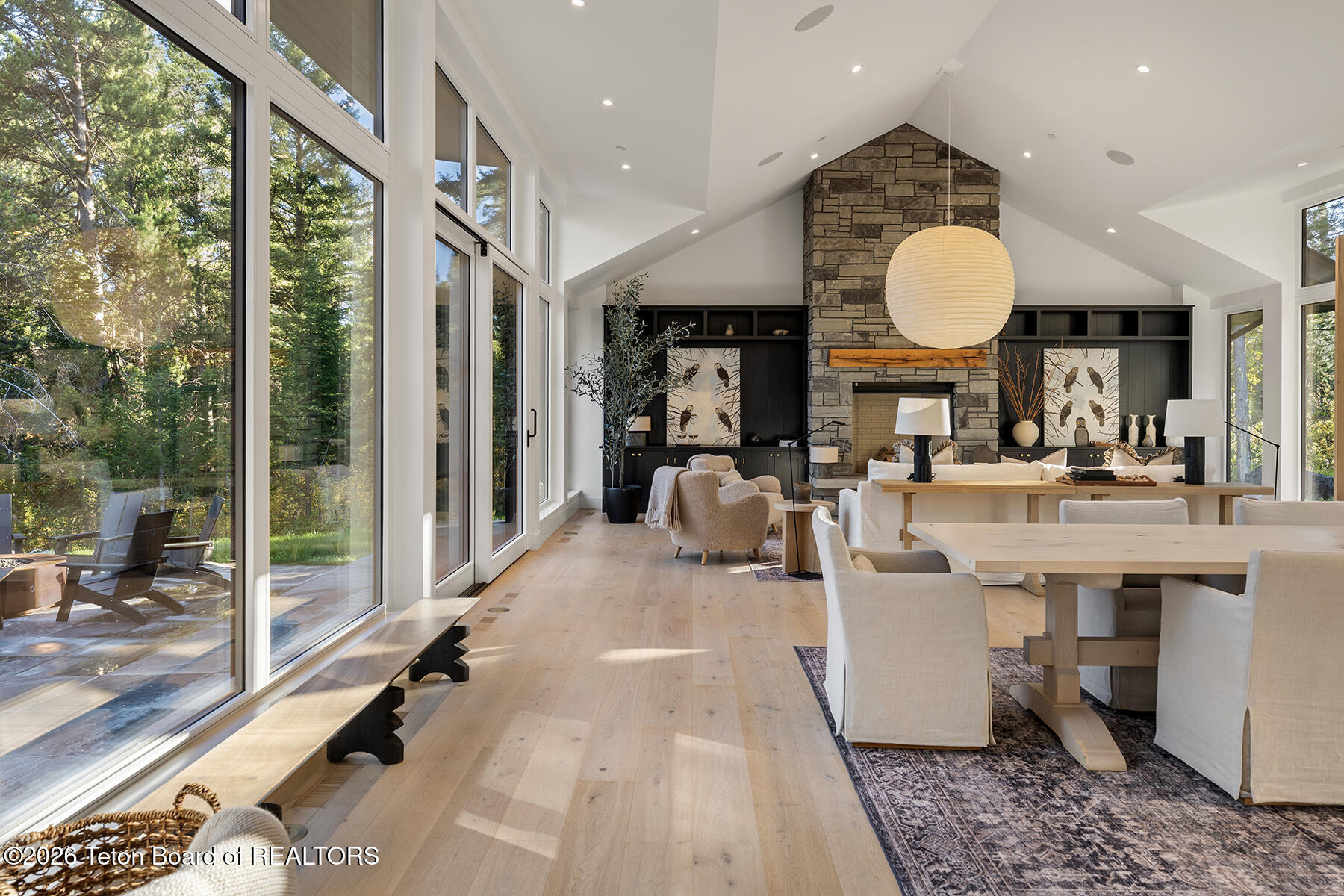 Open-plan living and dining room with a vaulted ceiling, tall stone fireplace, and expansive windows overlooking trees.