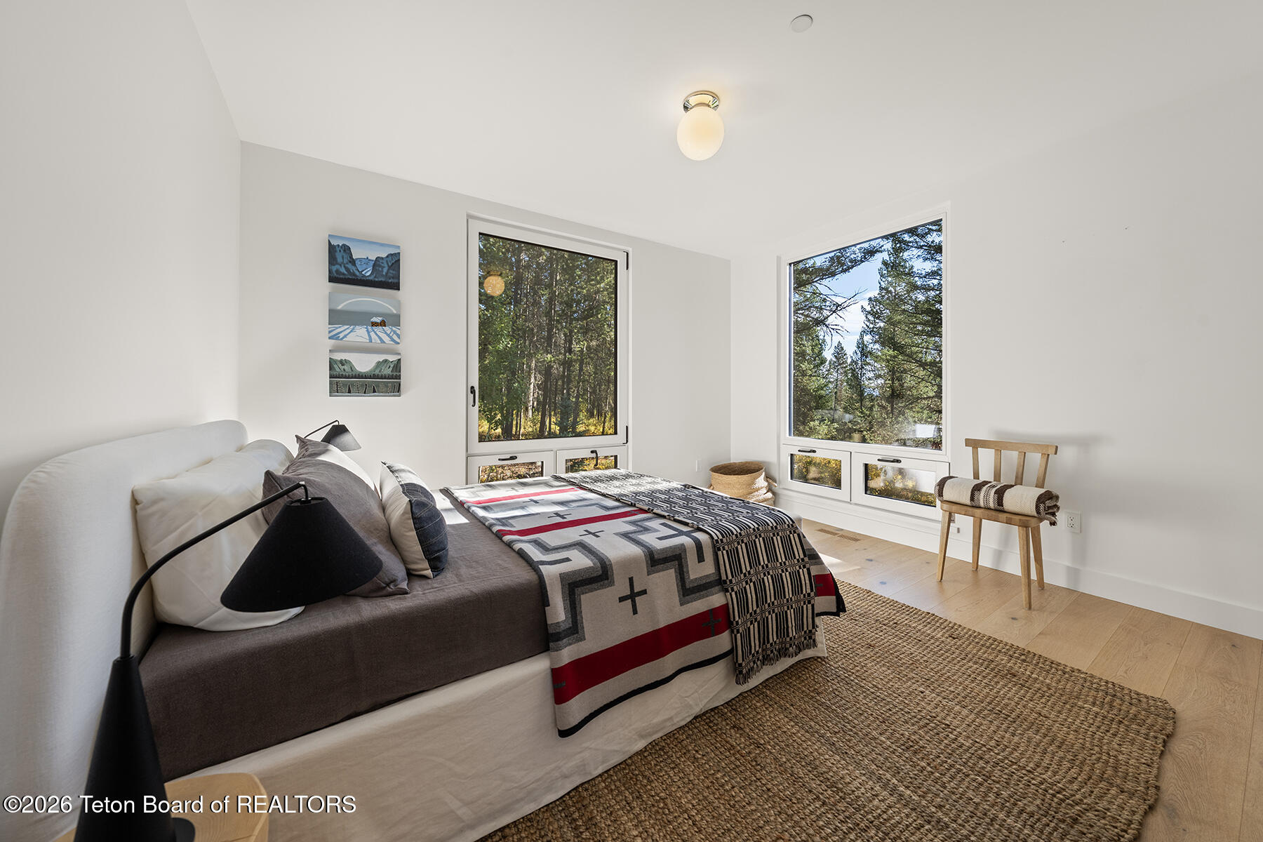 Furnished bedroom with large windows overlooking pine trees and a bed layered with geometric blankets.