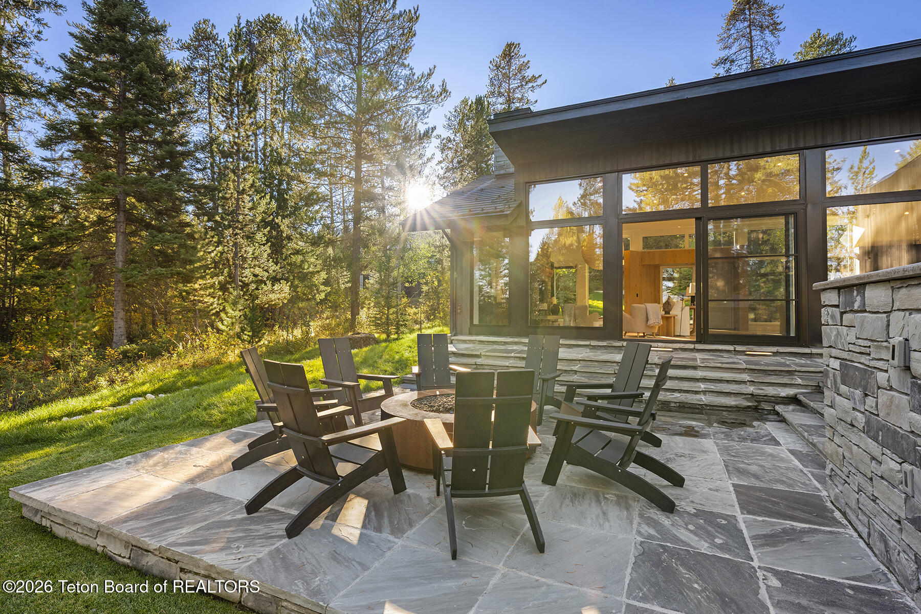 Modern home exterior with a stone patio, fire pit seating, and pine forest in golden-hour light.