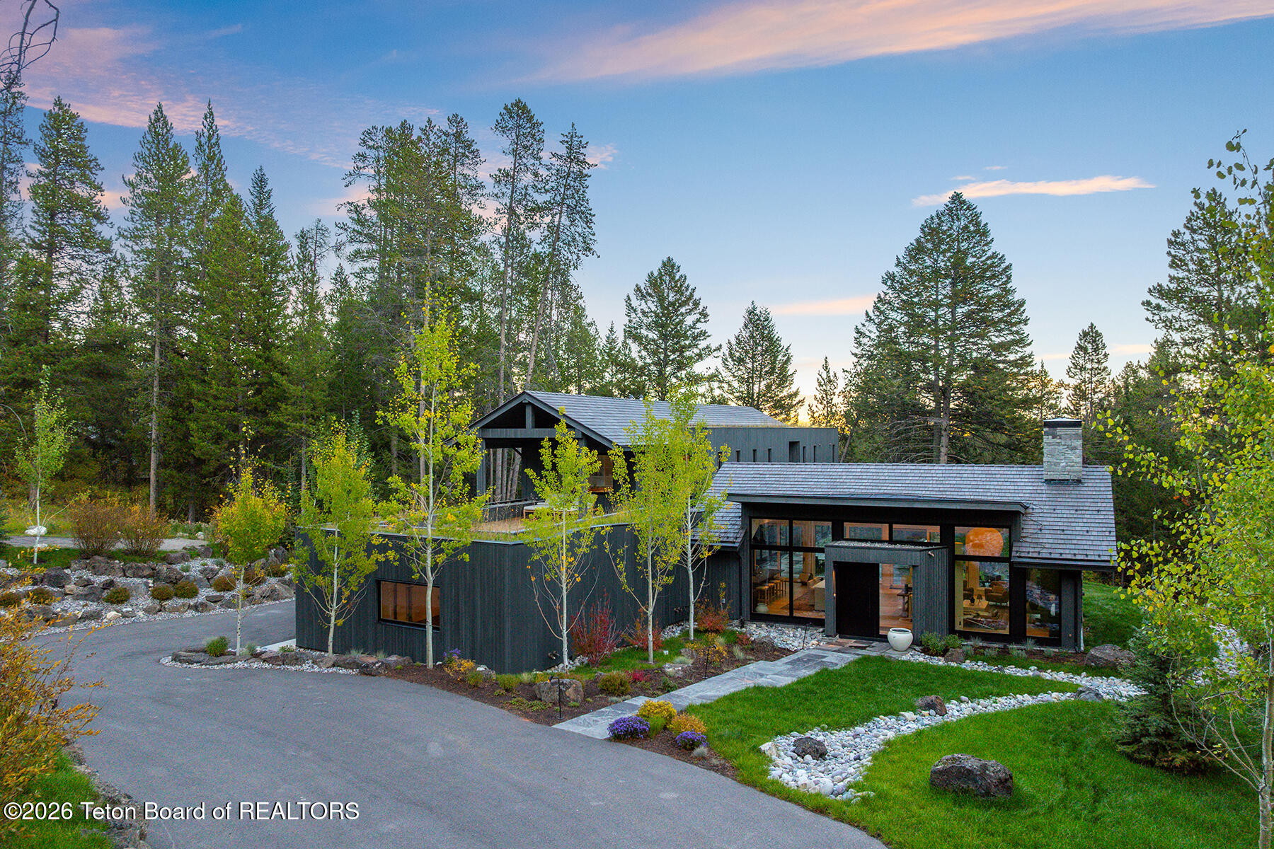 Modern home with broad gray roofs and large windows, surrounded by pine trees and landscaped gardens at dusk.