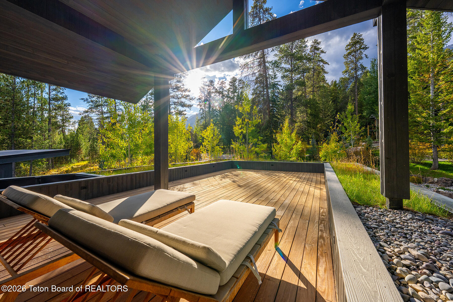 Covered wooden deck with chaise lounges overlooking sunlit trees and a distant mountain.