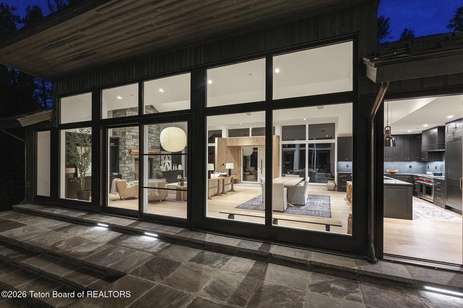 Modern home at dusk with floor-to-ceiling windows opening onto a stone patio.