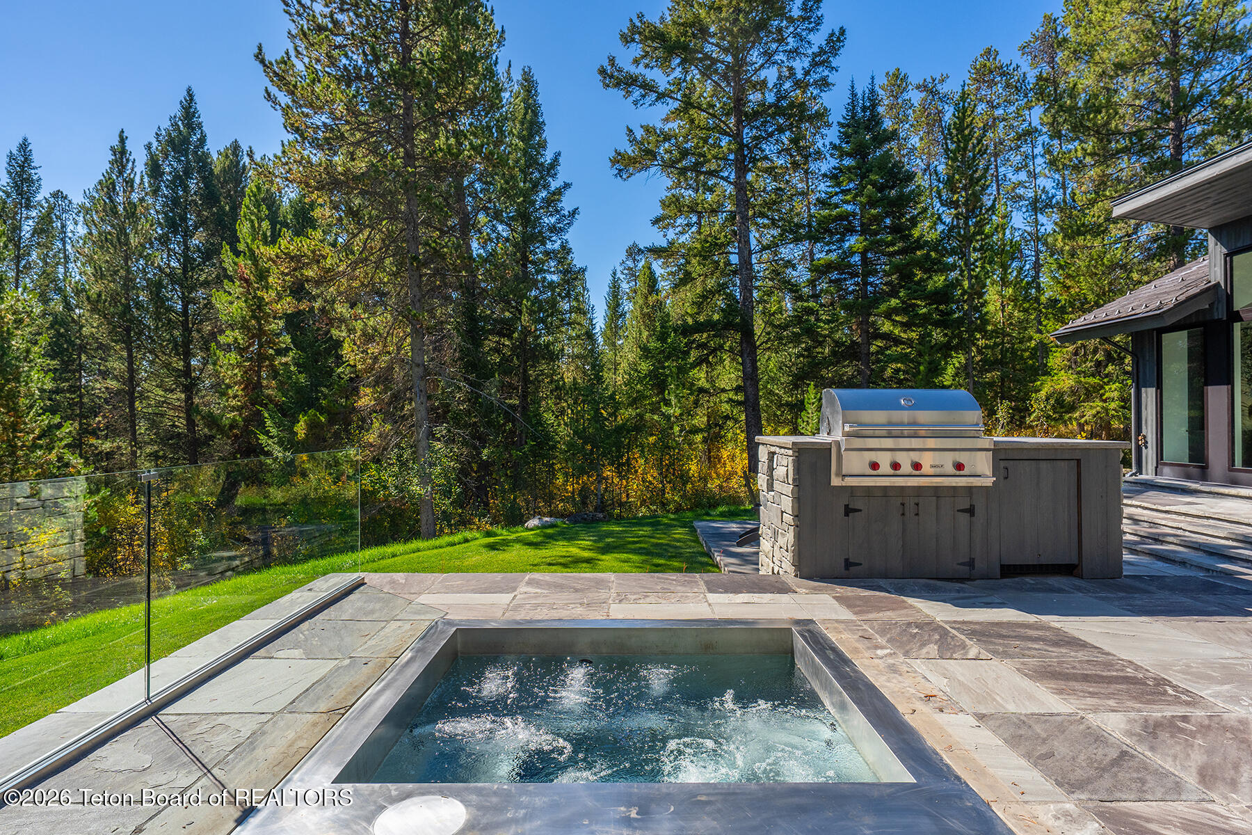 Stone patio with a plunge pool and built-in grill beside a wooded home.