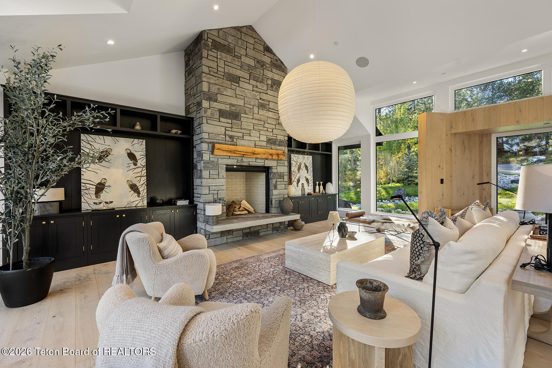 Living room with a tall stone fireplace, dark built-ins, and large windows overlooking landscaped grounds.