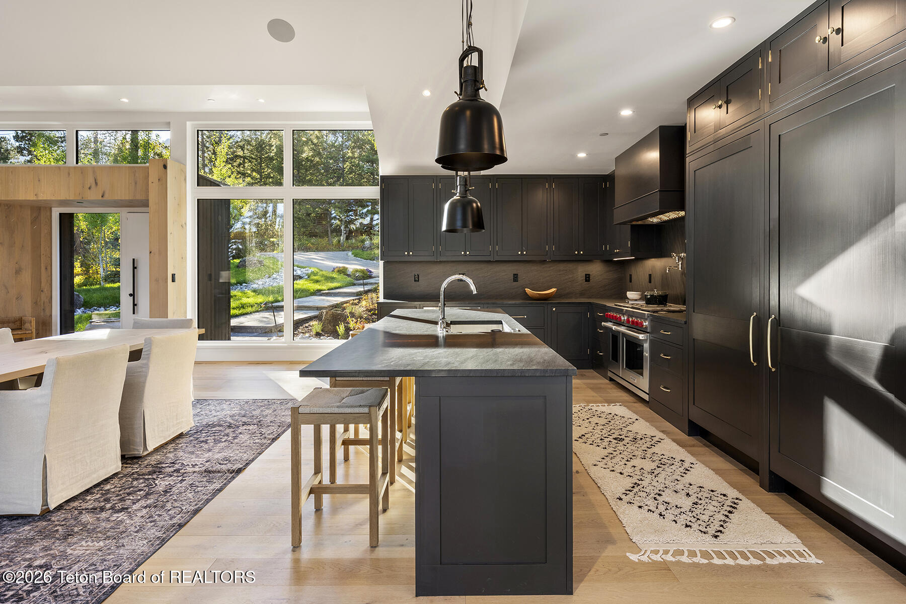 Open kitchen with a large stone island, dark cabinetry, and tall windows beside the dining area.