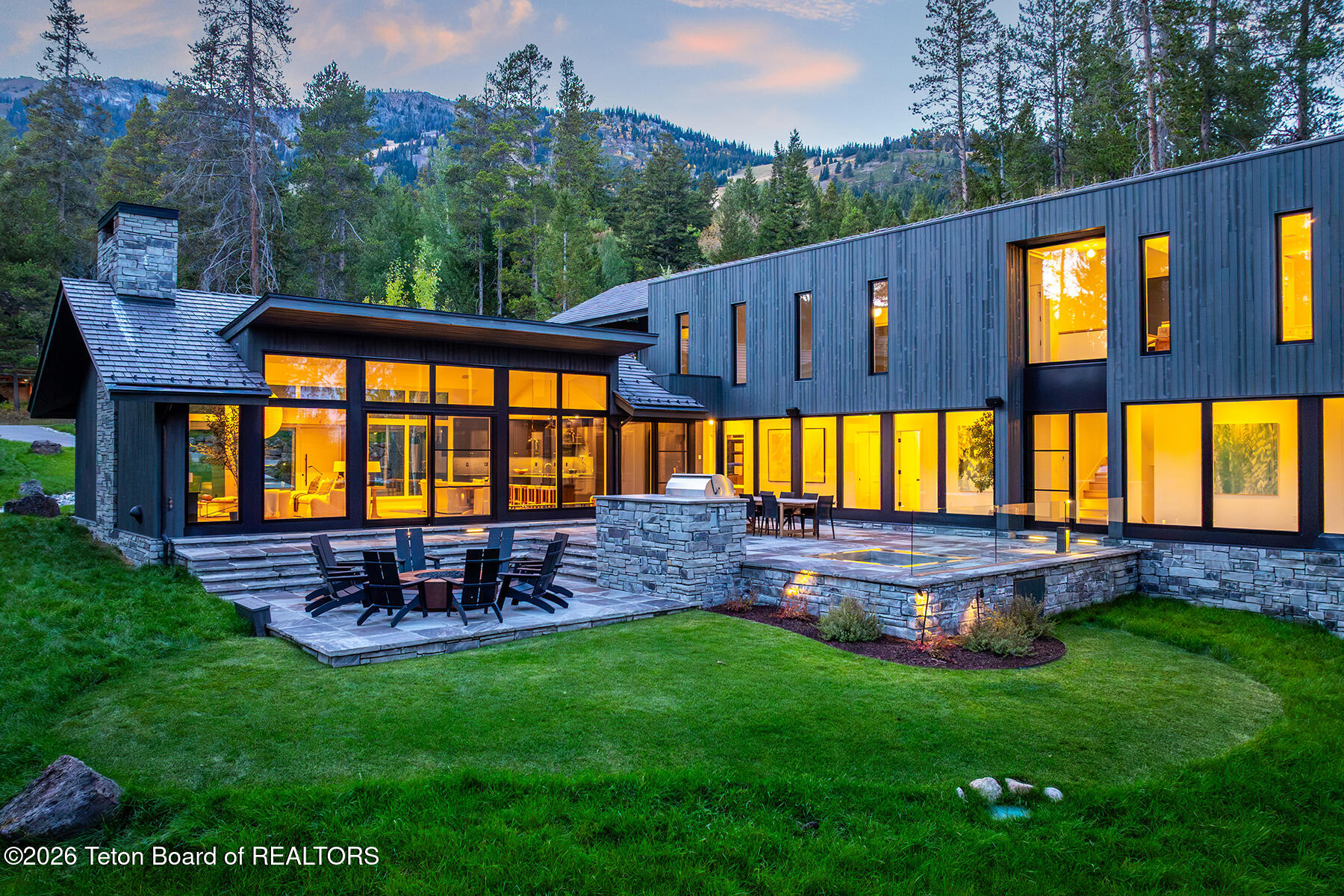 Modern mountain home with illuminated windows, stone patio, and outdoor dining area at dusk.