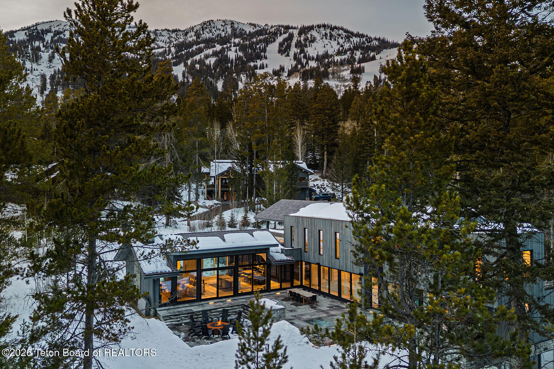 Modern glass-walled home in a snowy pine forest, with a mountain slope and illuminated interiors.
