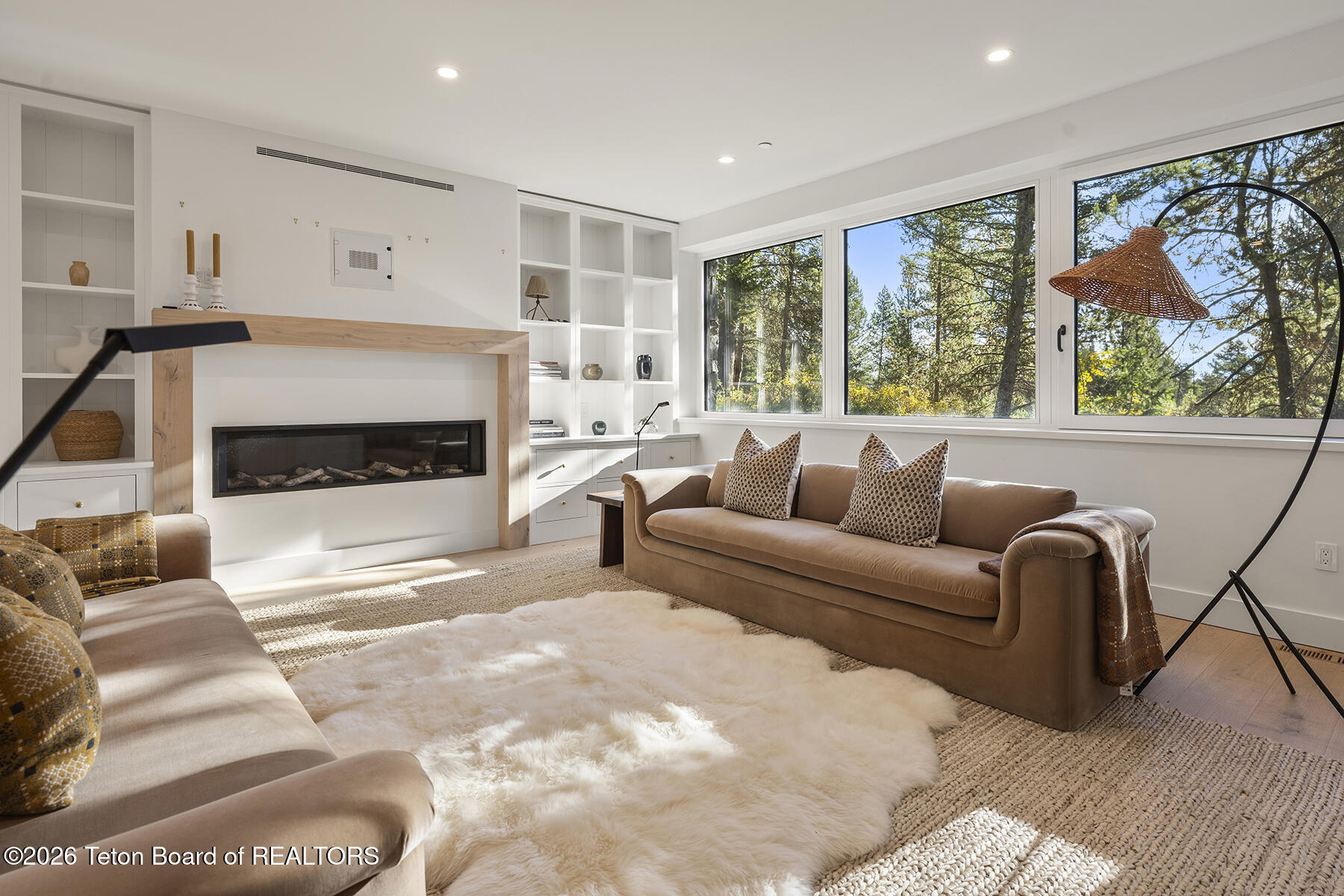 Living room with a linear fireplace, built-in shelving, and large windows overlooking trees.