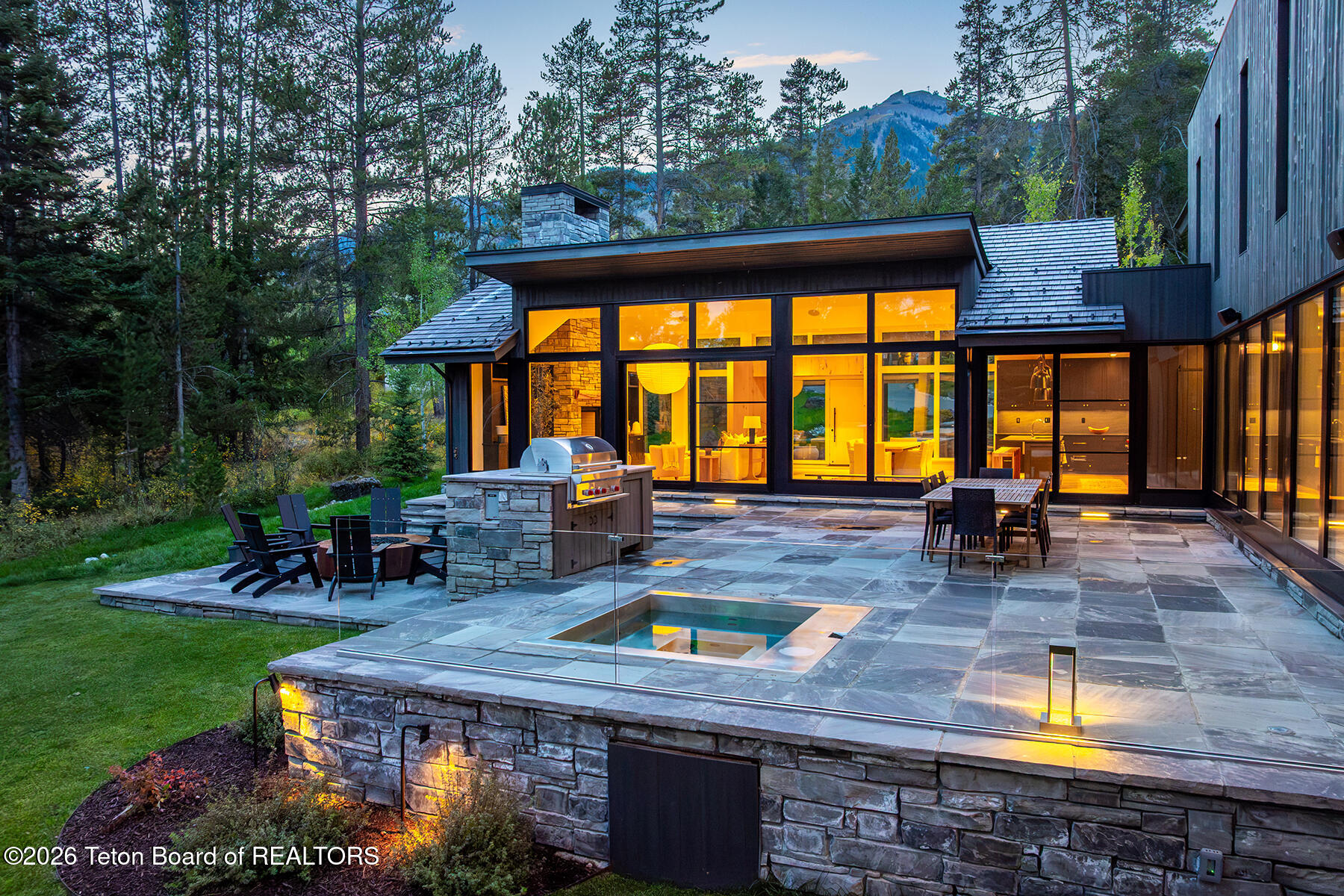 Modern mountain home with illuminated glass walls, stone patio, outdoor kitchen, and plunge pool.
