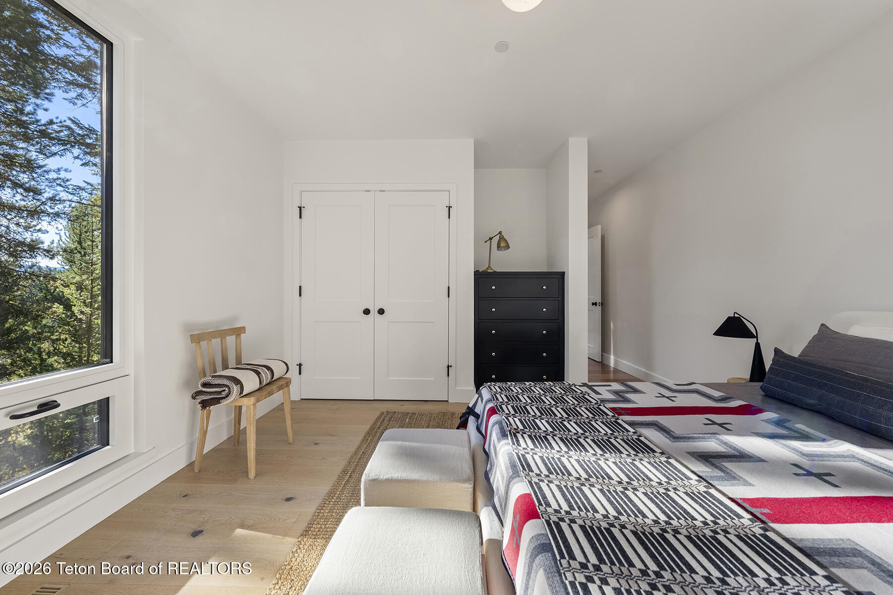 Furnished bedroom with a patterned bed and a large window overlooking evergreen trees.