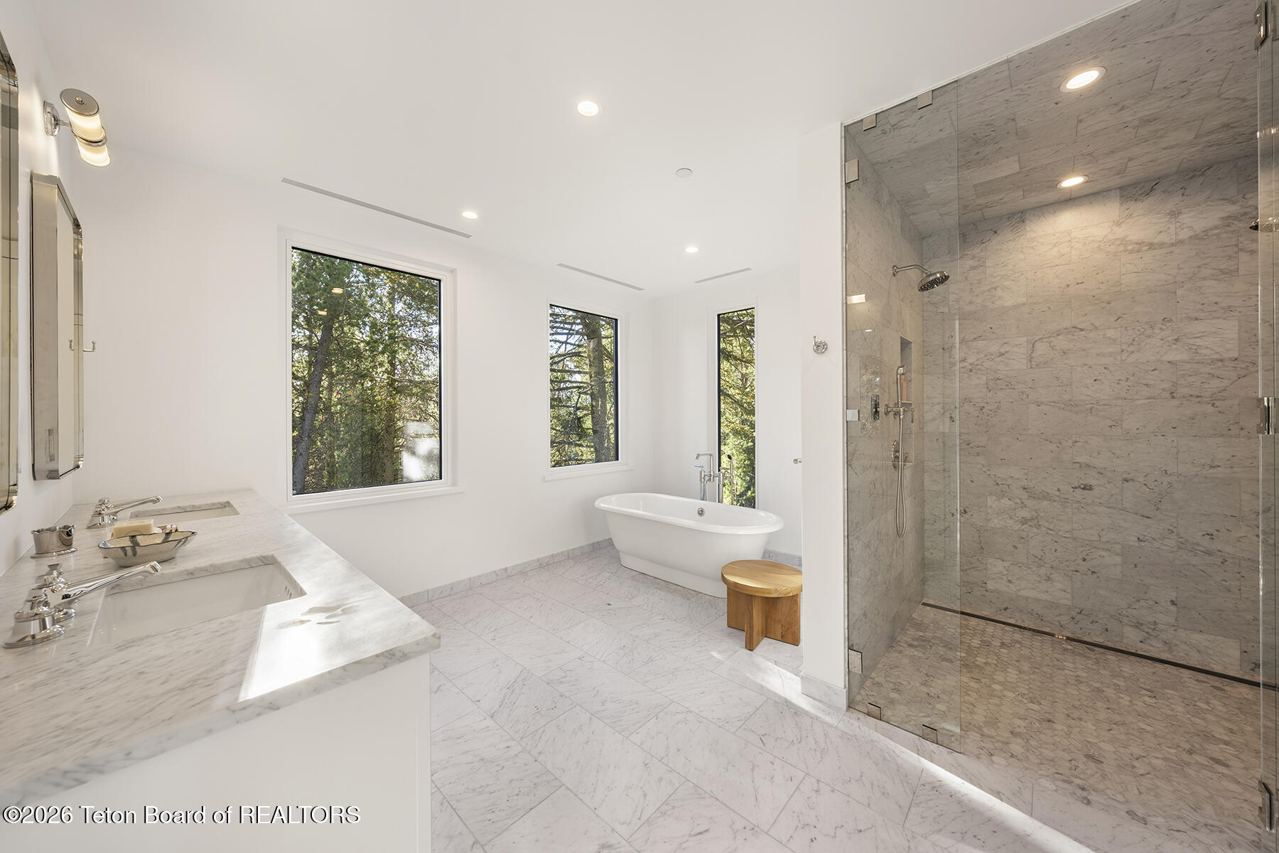 Marble bathroom with a freestanding tub, glass shower, double vanity, and windows overlooking trees.