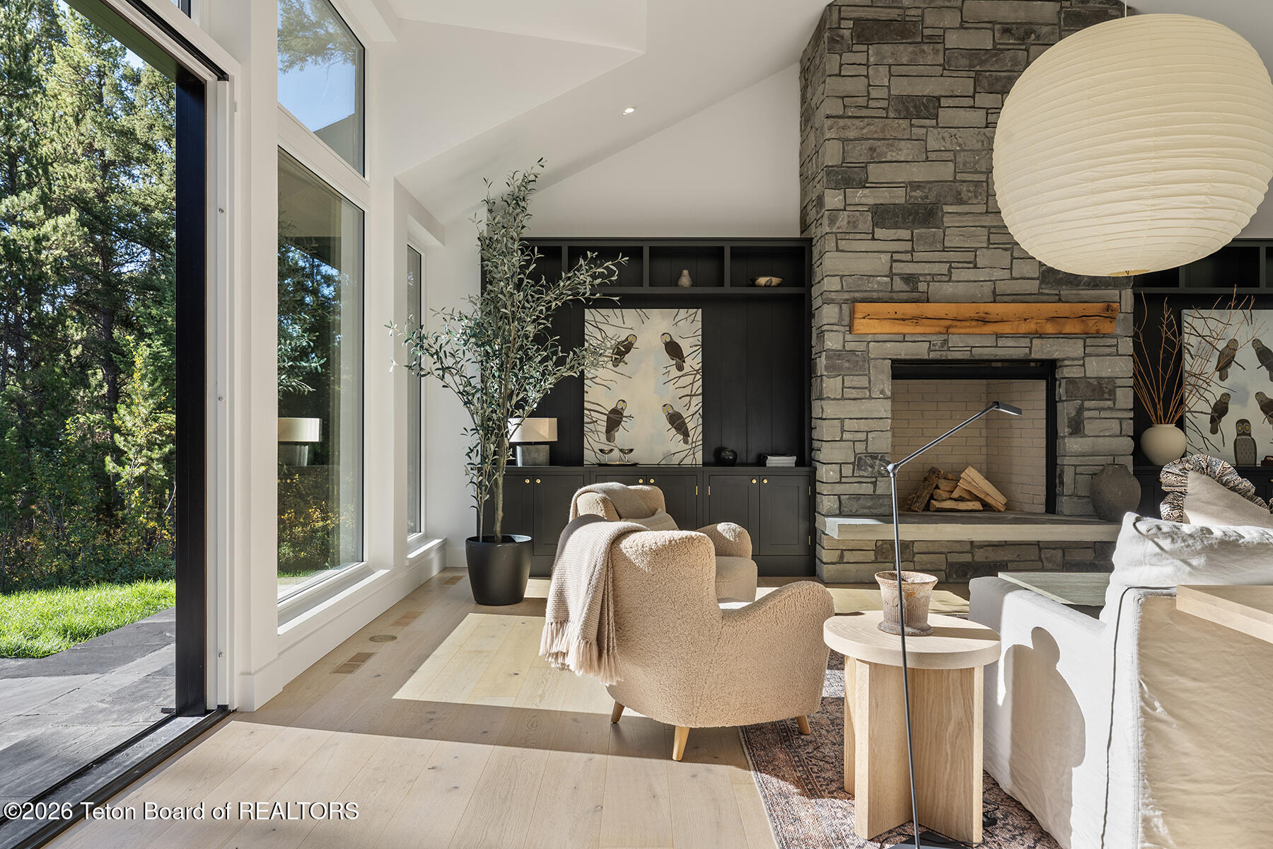 Living room with a stone fireplace, dark built-ins, and large windows overlooking trees.