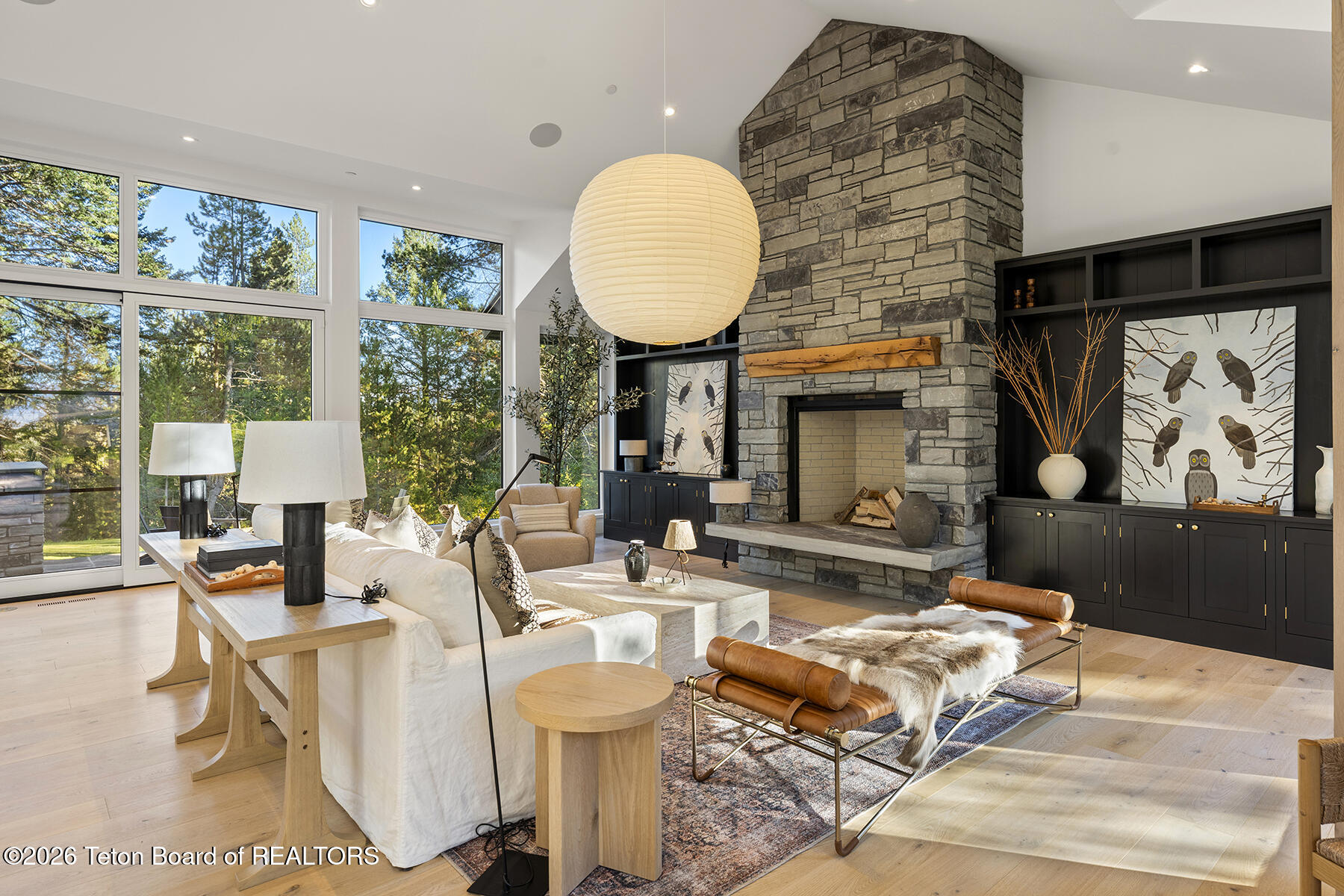 Staged living room with a tall stone fireplace, dark built-ins, and large windows overlooking trees.