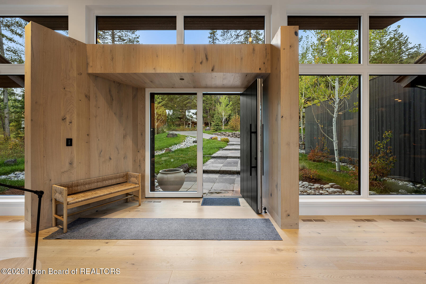 Modern wood-paneled entry foyer with a built-in bench, tall windows, and an open door to the garden.
