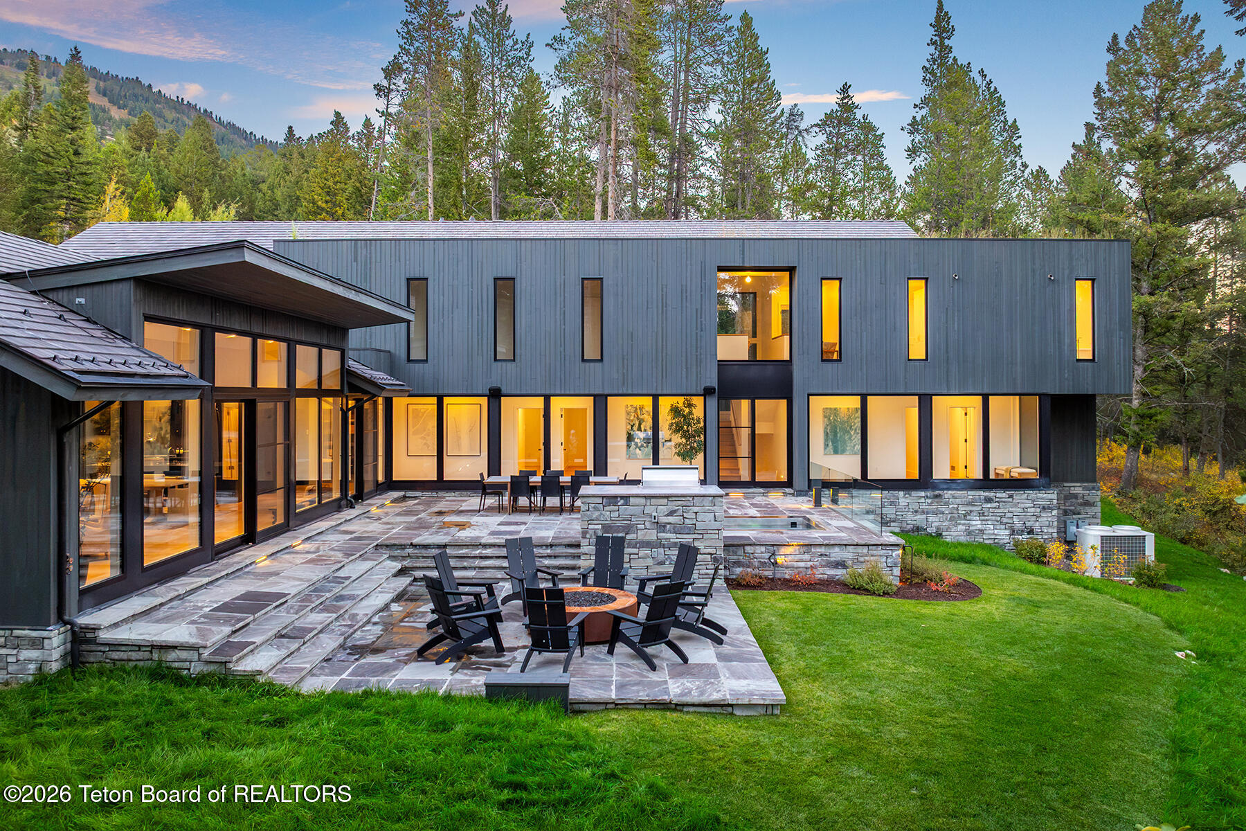 Modern home with stone patio and fire pit, backed by pine forest and mountain slope.
