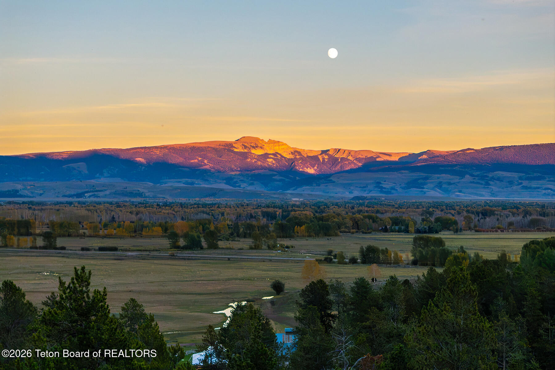 Mountain range glowing above a rural valley at golden hour, with the moon visible overhead.
