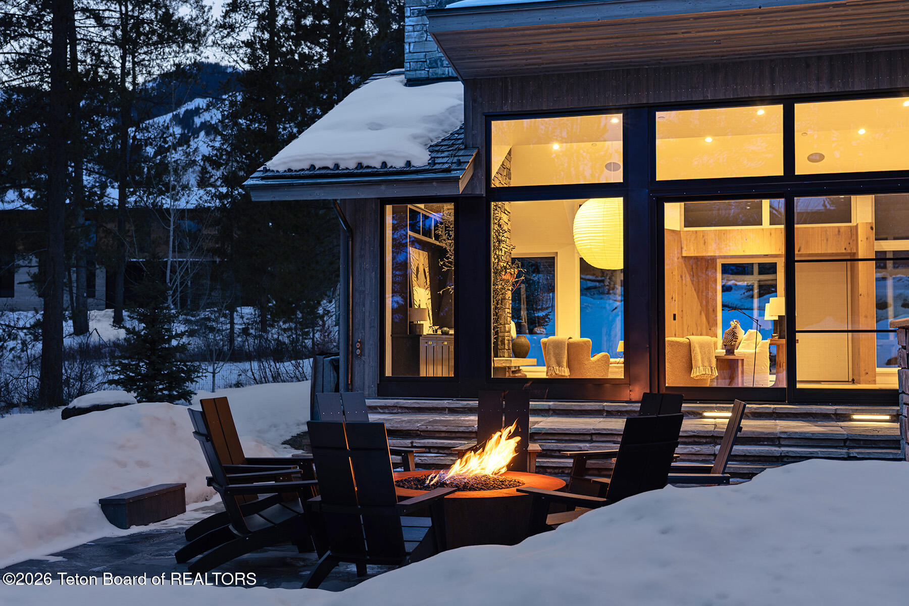 Modern glass-fronted home glowing at dusk above a snowy patio fire pit.