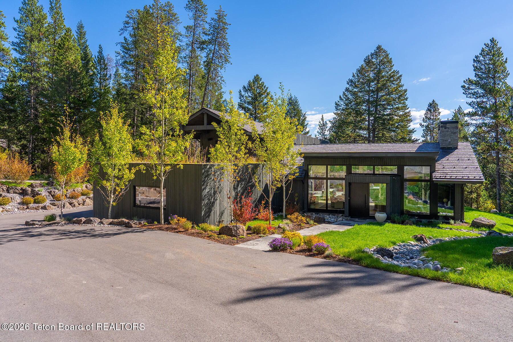 Contemporary dark-sided home with broad windows and stone chimney among evergreens and autumn trees.