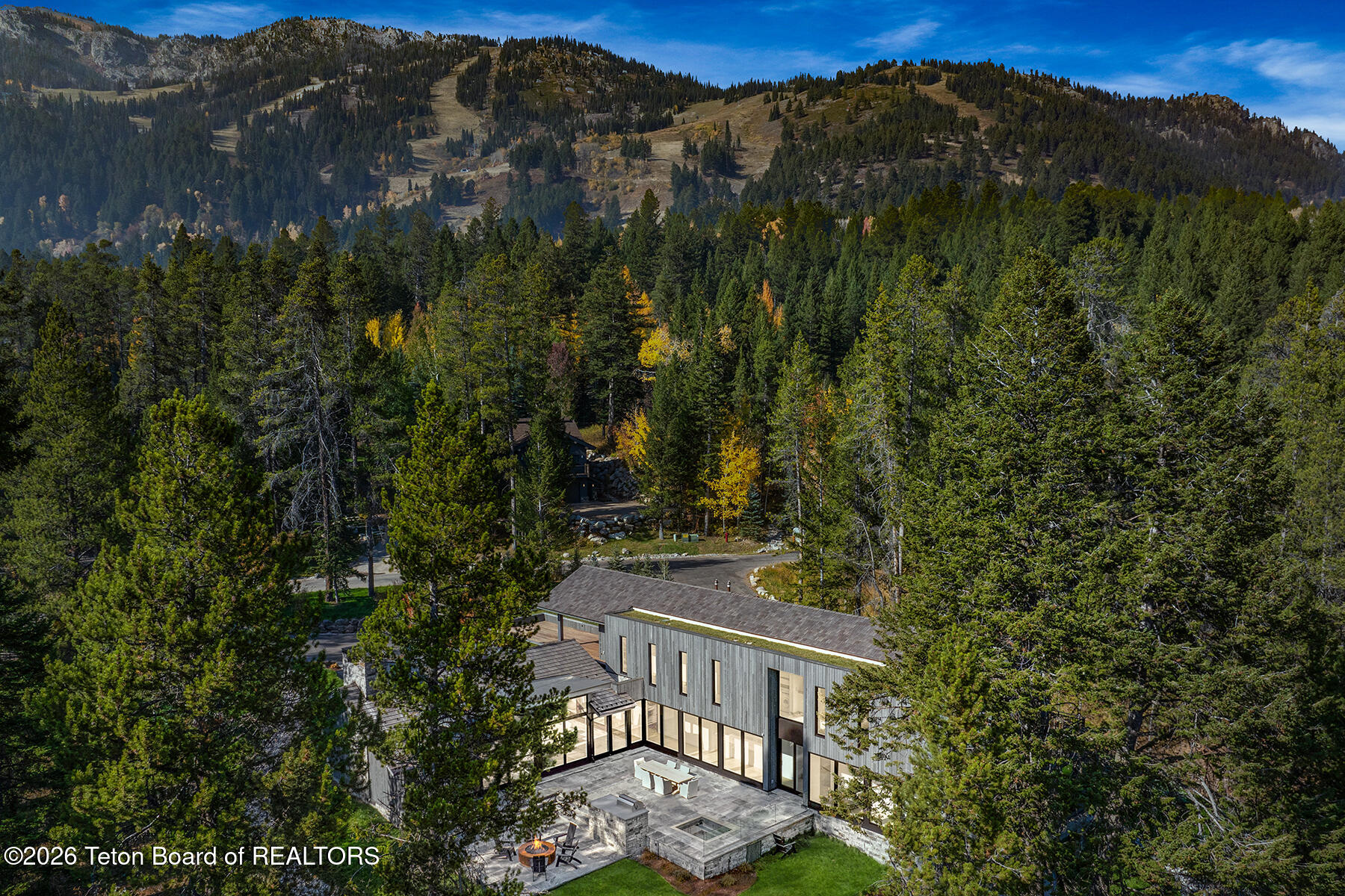 A modern home surrounded by forest, with a stone terrace and mountain ridges in the background.