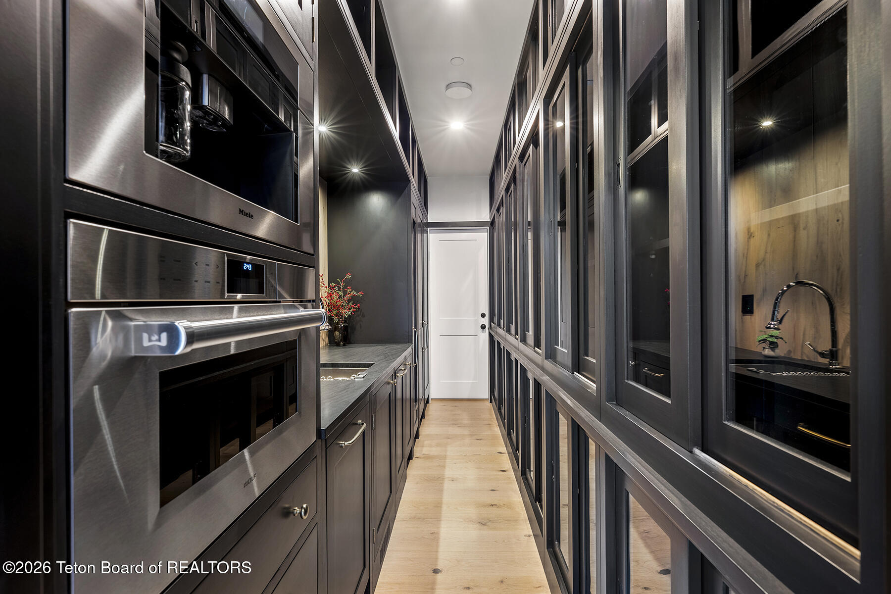 Modern galley kitchen pantry with dark glass-front cabinets, built-in stainless steel ovens, and pale wood flooring.