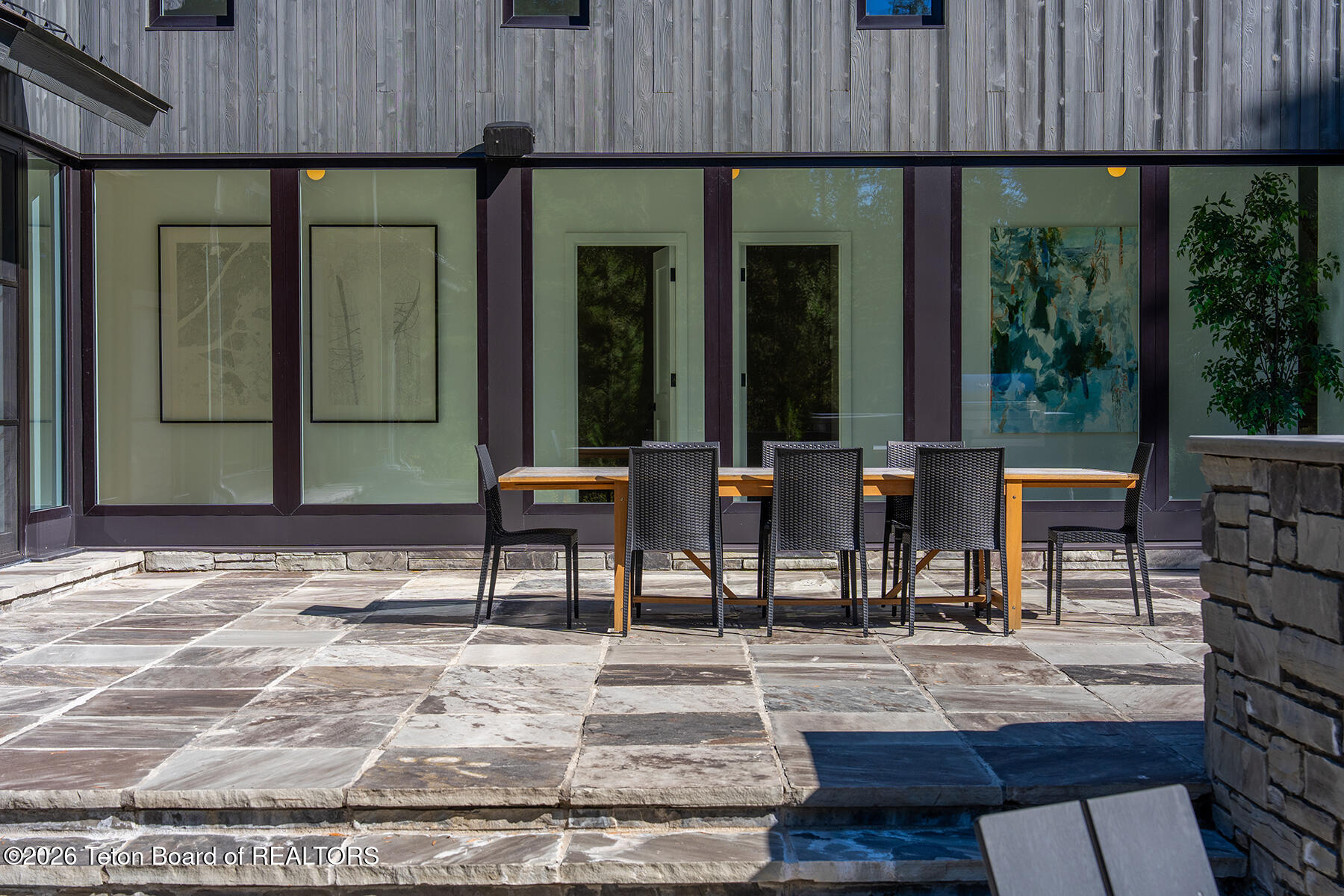 Stone patio with outdoor dining table and chairs beside modern glass walls, next to a pool.