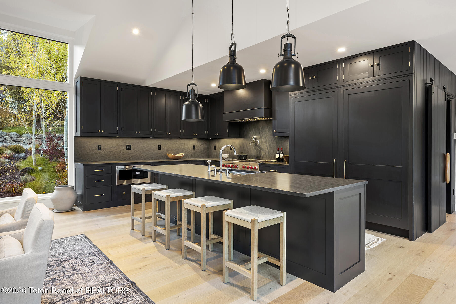Modern kitchen with black cabinetry, a large island, pendant lights, and tall windows overlooking autumn foliage.