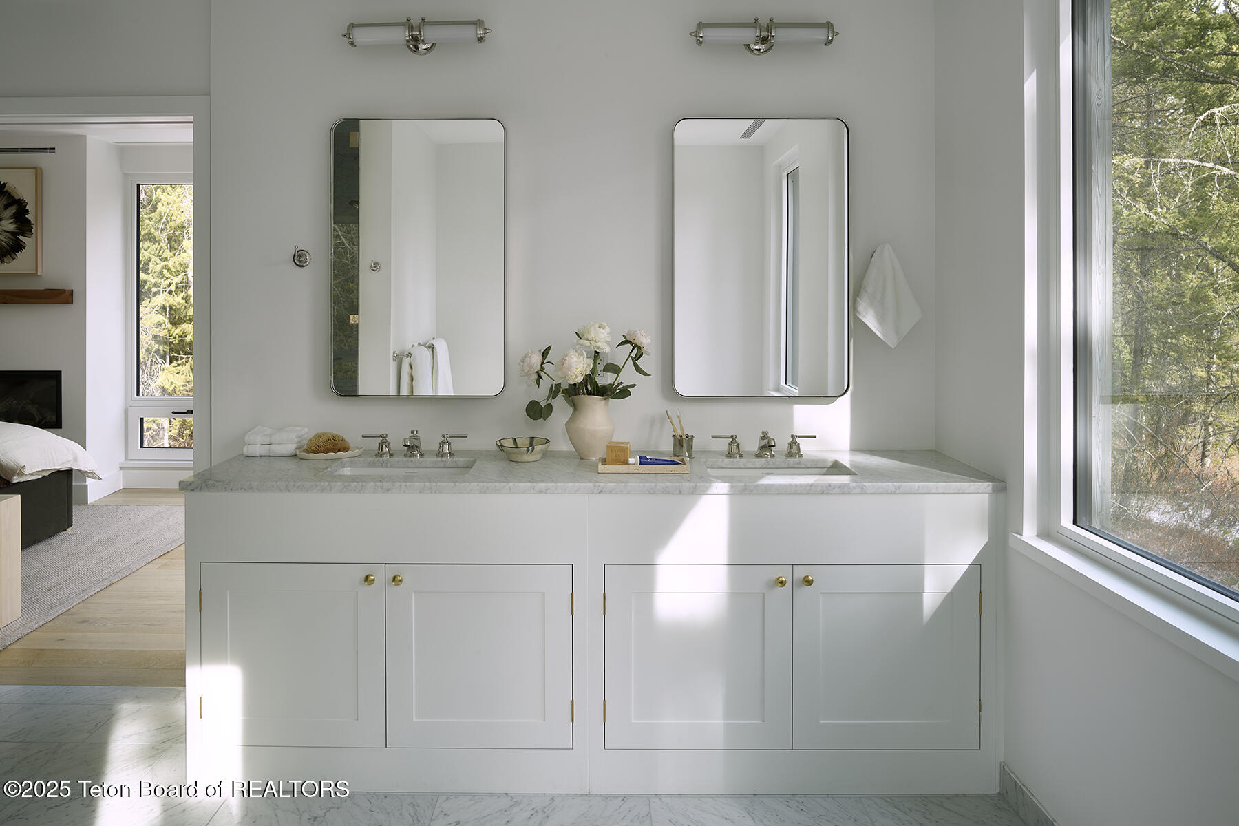 Bright bathroom with a double vanity, marble counter, brass fixtures, and large windows overlooking trees.