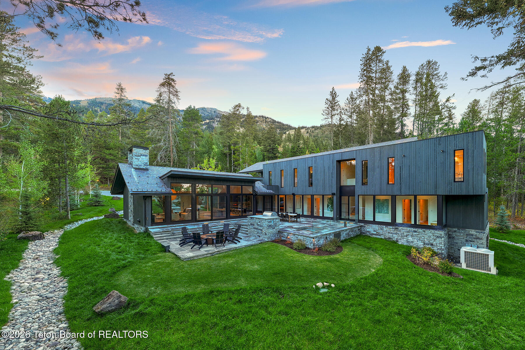 Modern mountain home with a stone patio, green lawn, and forested mountain backdrop.