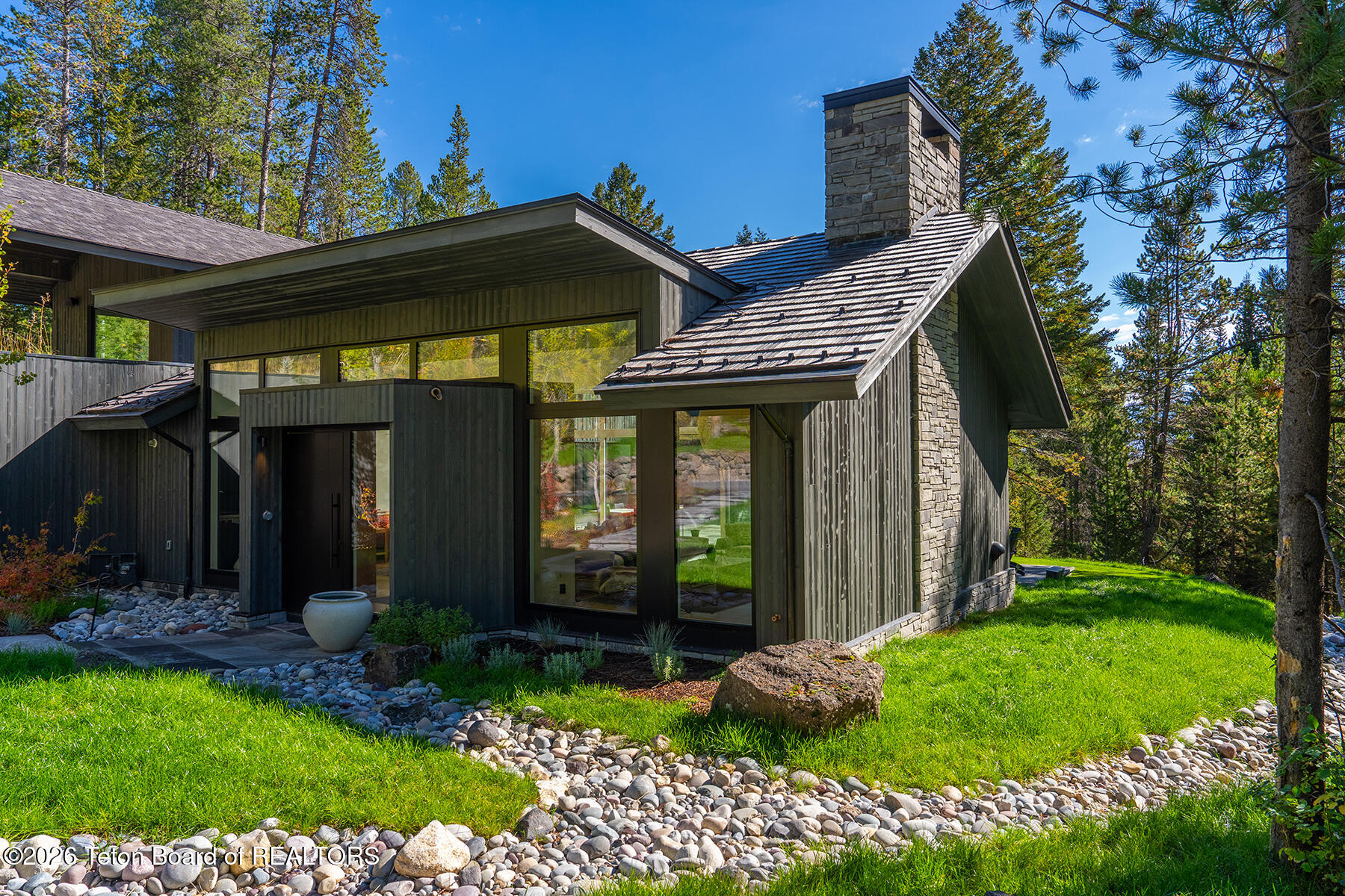 Modern wood-sided home with a stone chimney, broad windows, and pine trees.