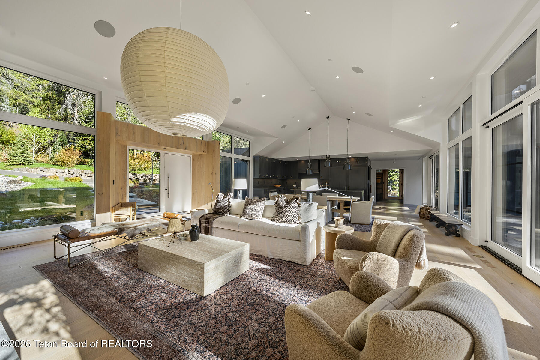 Open-plan living room with vaulted ceiling, oversized pendant light, and expansive windows overlooking landscaped grounds.