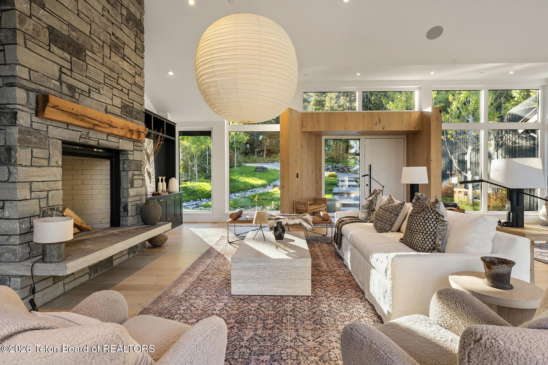 Living room with a stone fireplace, white sofas, and large windows overlooking a landscaped yard.