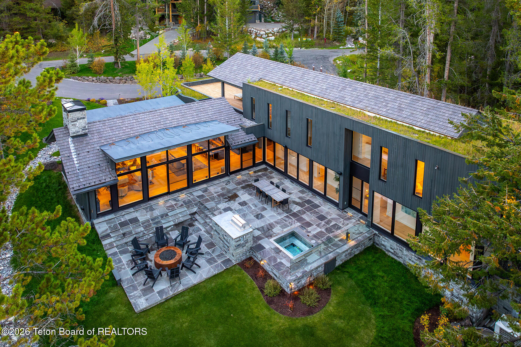 Modern home with a stone patio, fire pit, and small spa, viewed from above among autumn trees.