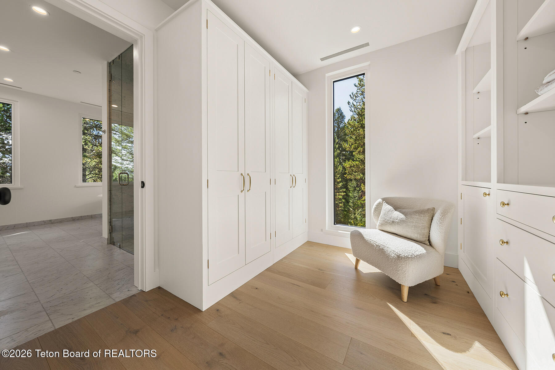 Walk-in closet with white built-ins, an upholstered chair, and a tall window overlooking trees.