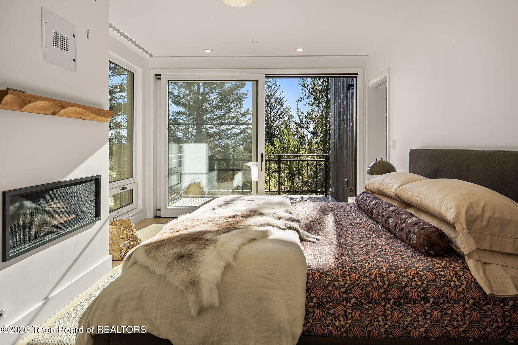 Bedroom with a wall fireplace and sliding doors opening to a tree-surrounded balcony.