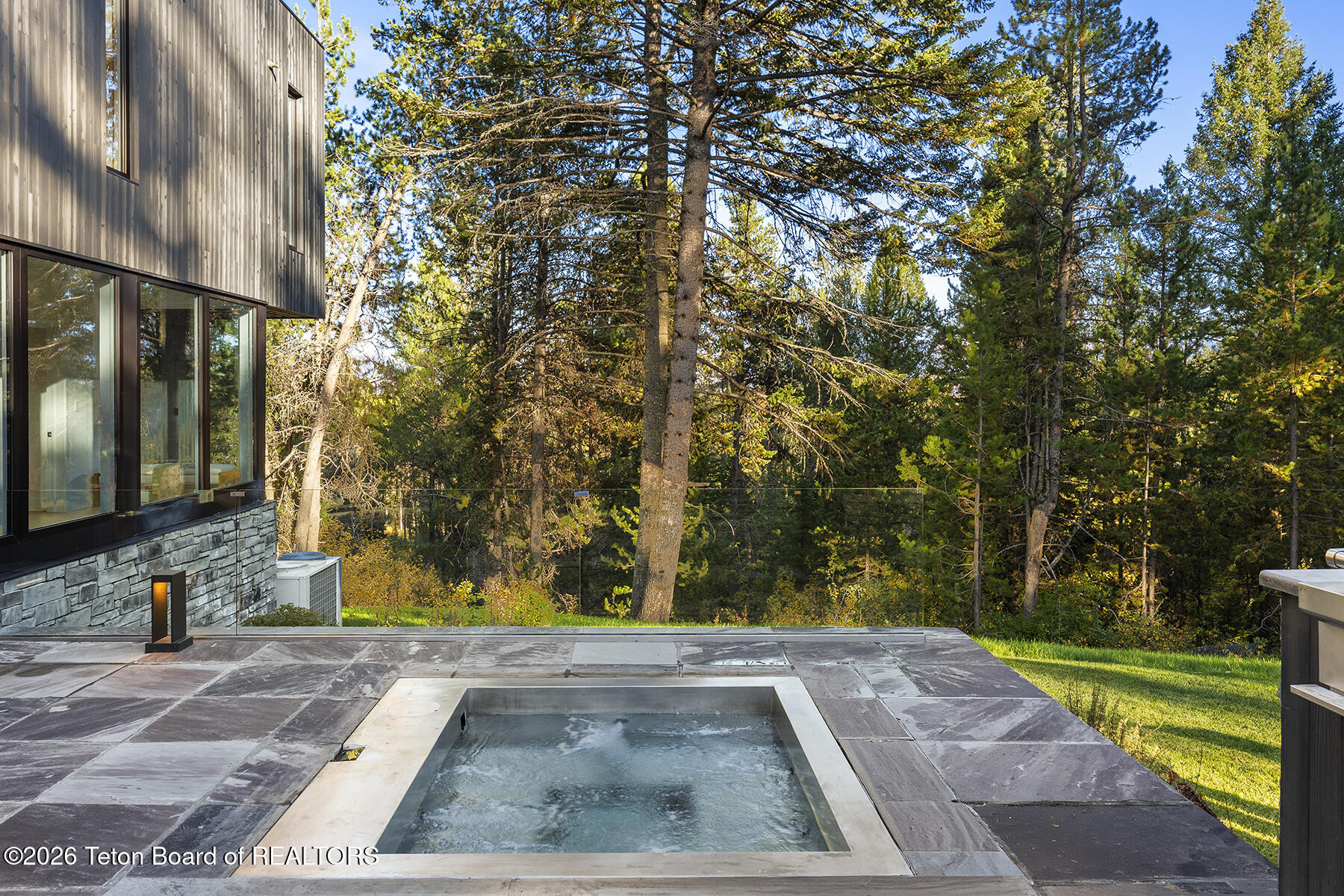 Stone patio spa beside a modern home, with glass railing and pine trees.