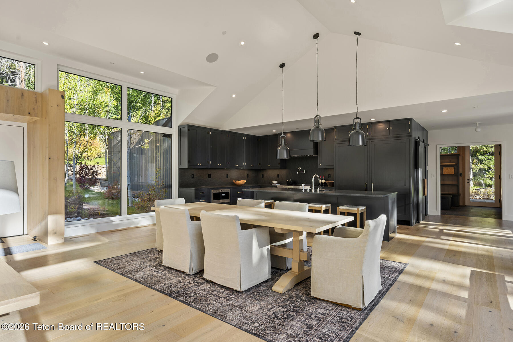Open-plan kitchen and dining area with dark cabinetry, a large island, and tall windows overlooking trees.