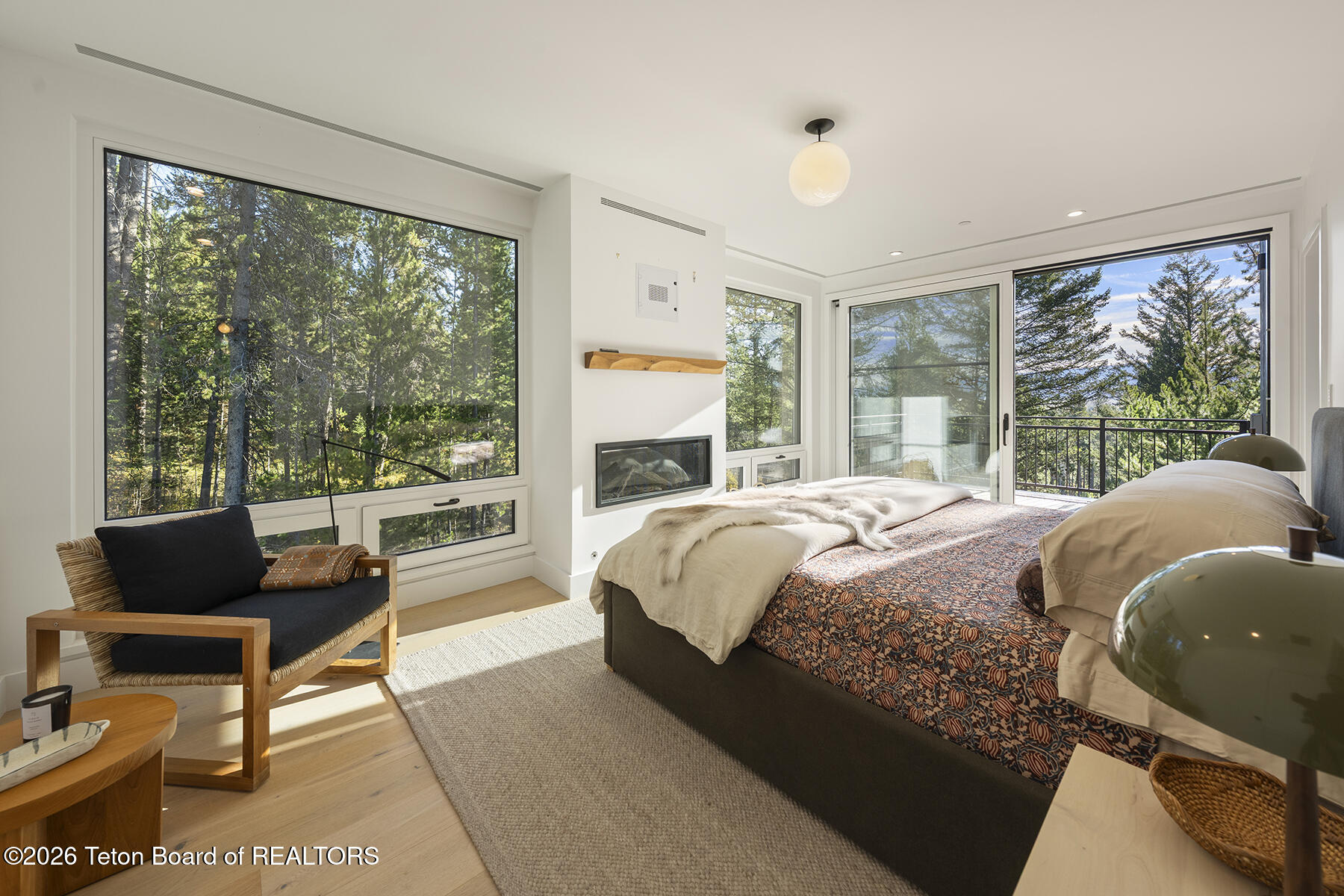 Modern bedroom with a built-in fireplace and floor-to-ceiling windows overlooking trees and distant mountains.
