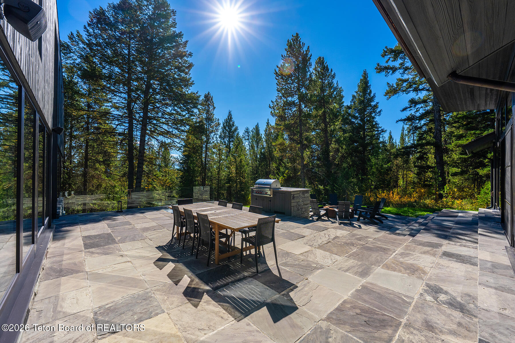 Stone patio with outdoor dining set, built-in grill, and seating beside a forest.