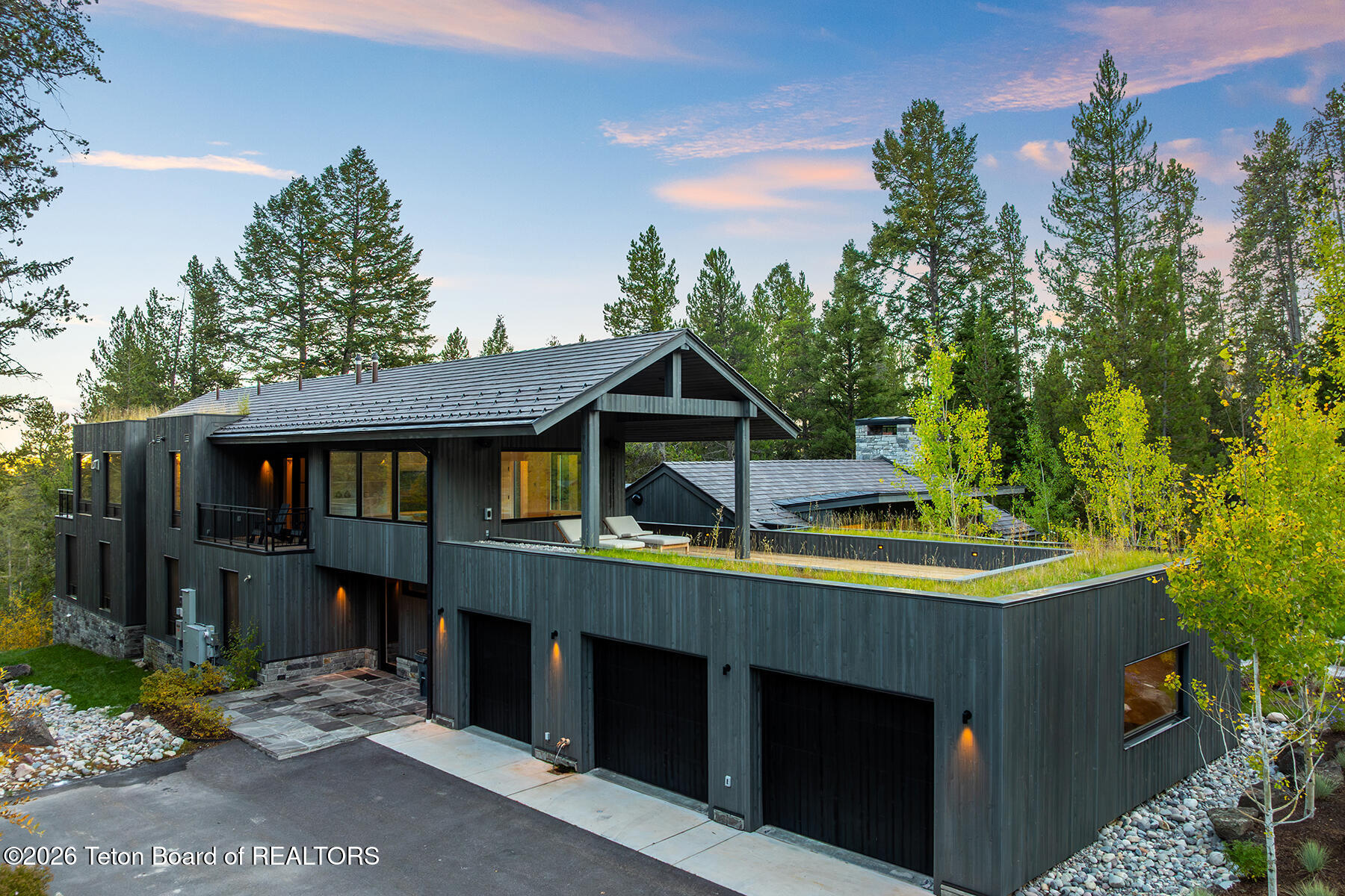 Modern dark-sided home with a raised pool, multiple garage doors, and autumn trees at dusk.