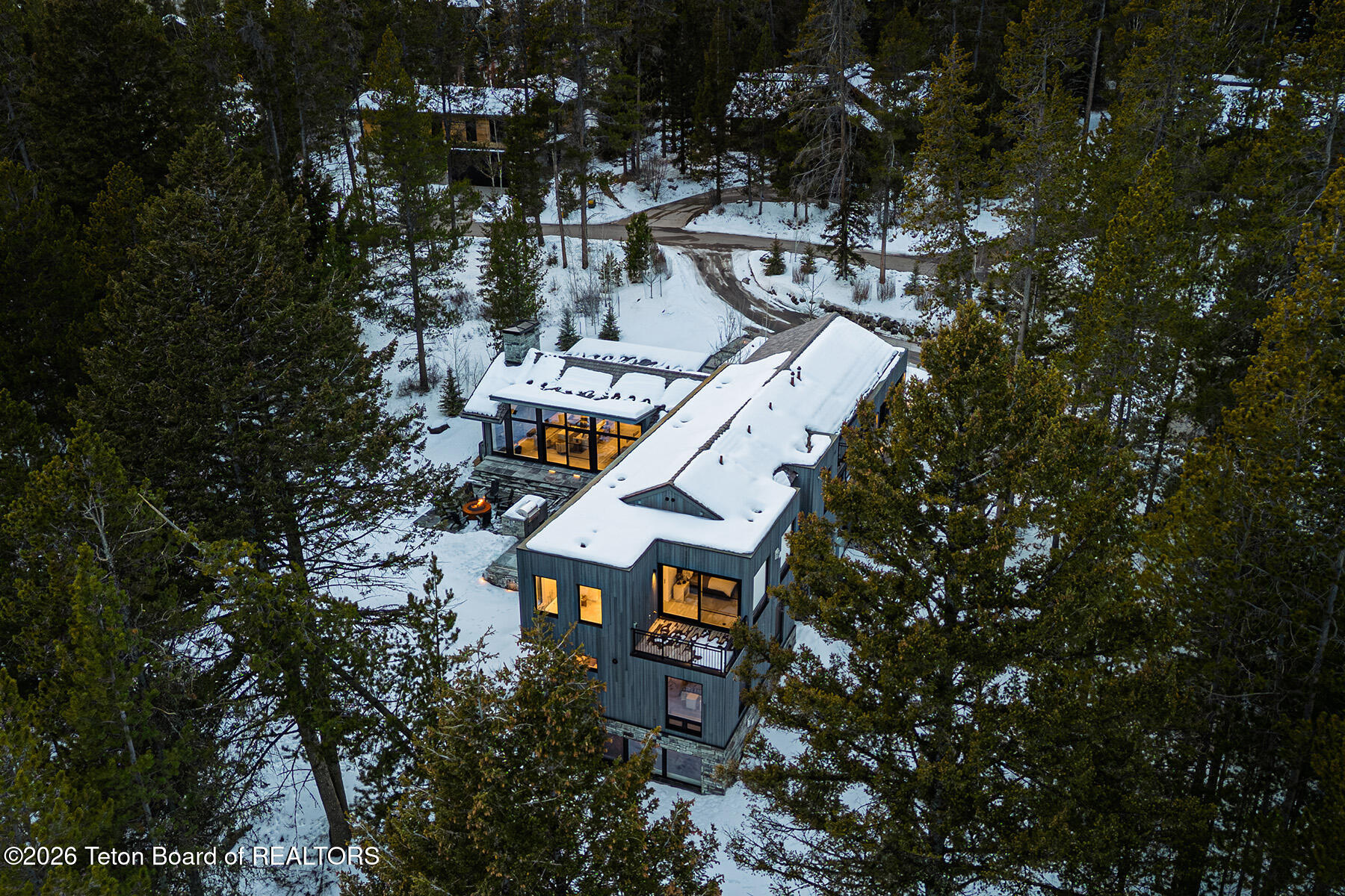 Modern forest home with a snow-covered roof and glowing windows, surrounded by evergreens.