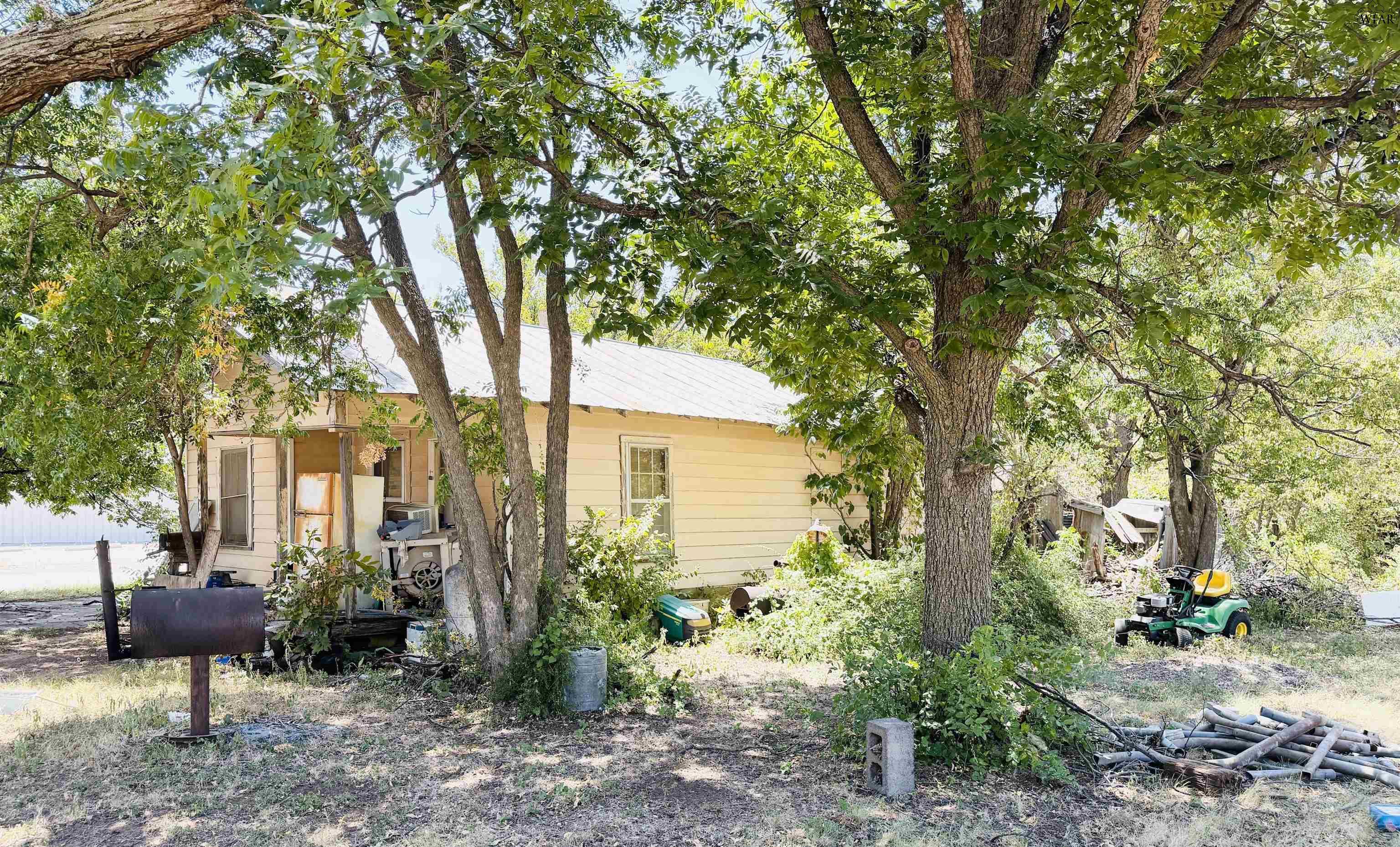 Pale yellow house partly hidden by mature trees in a cluttered yard.