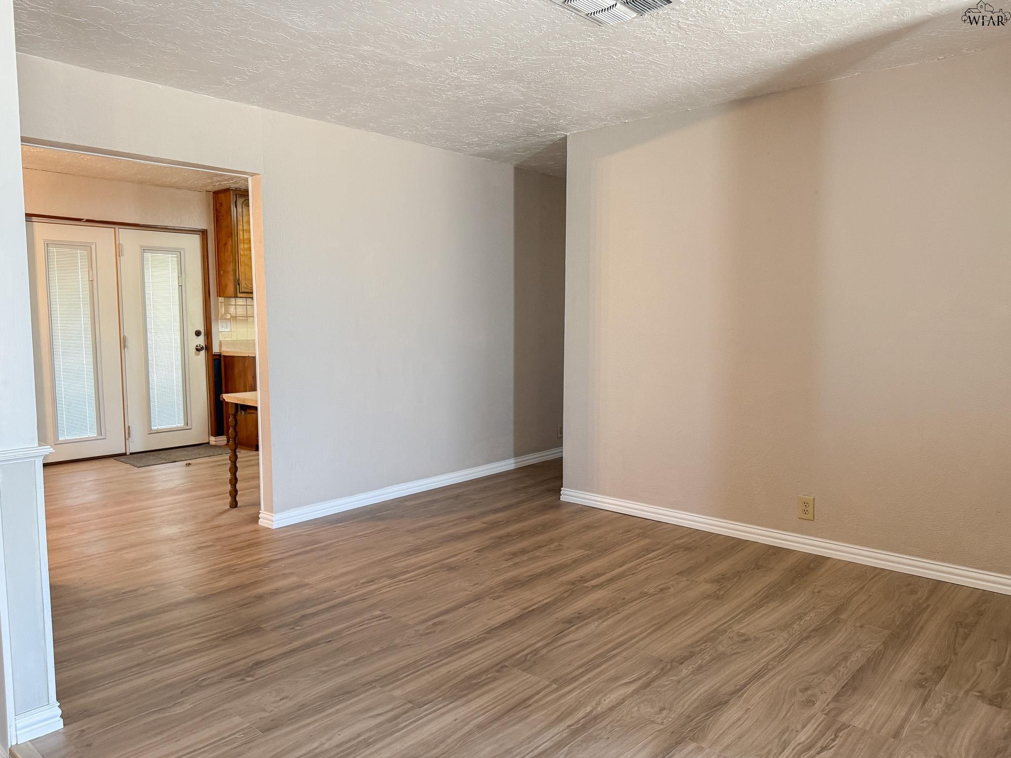 Empty living area with wood-look flooring and an opening to the kitchen and double doors.