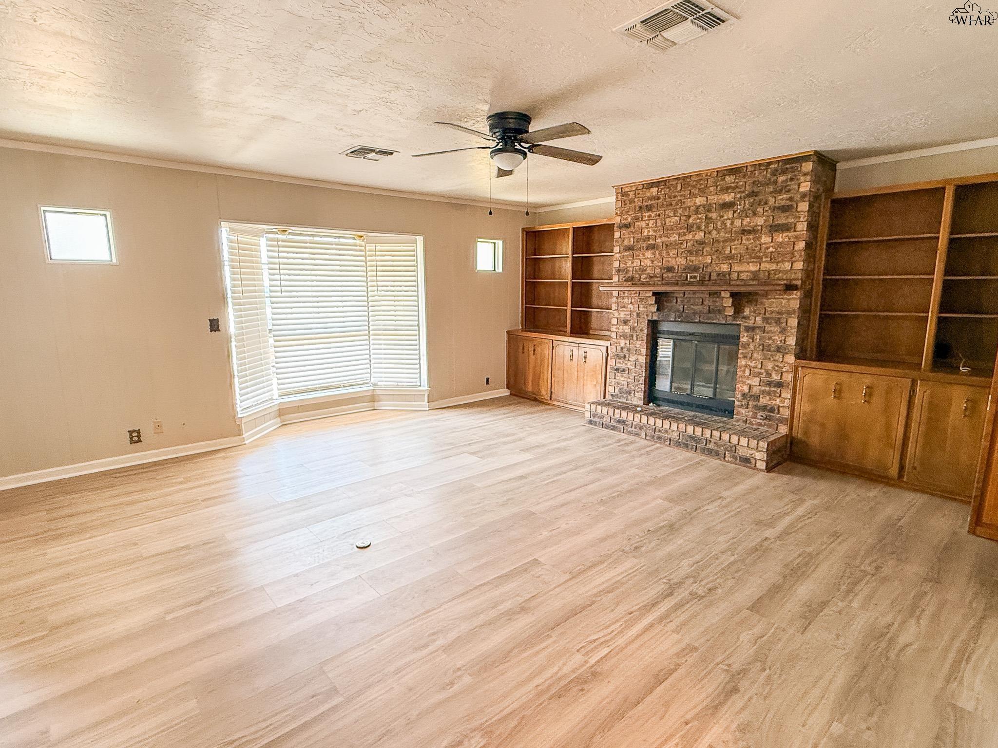Vacant living room with a brick fireplace, built-in shelving, and a large bay window with blinds.