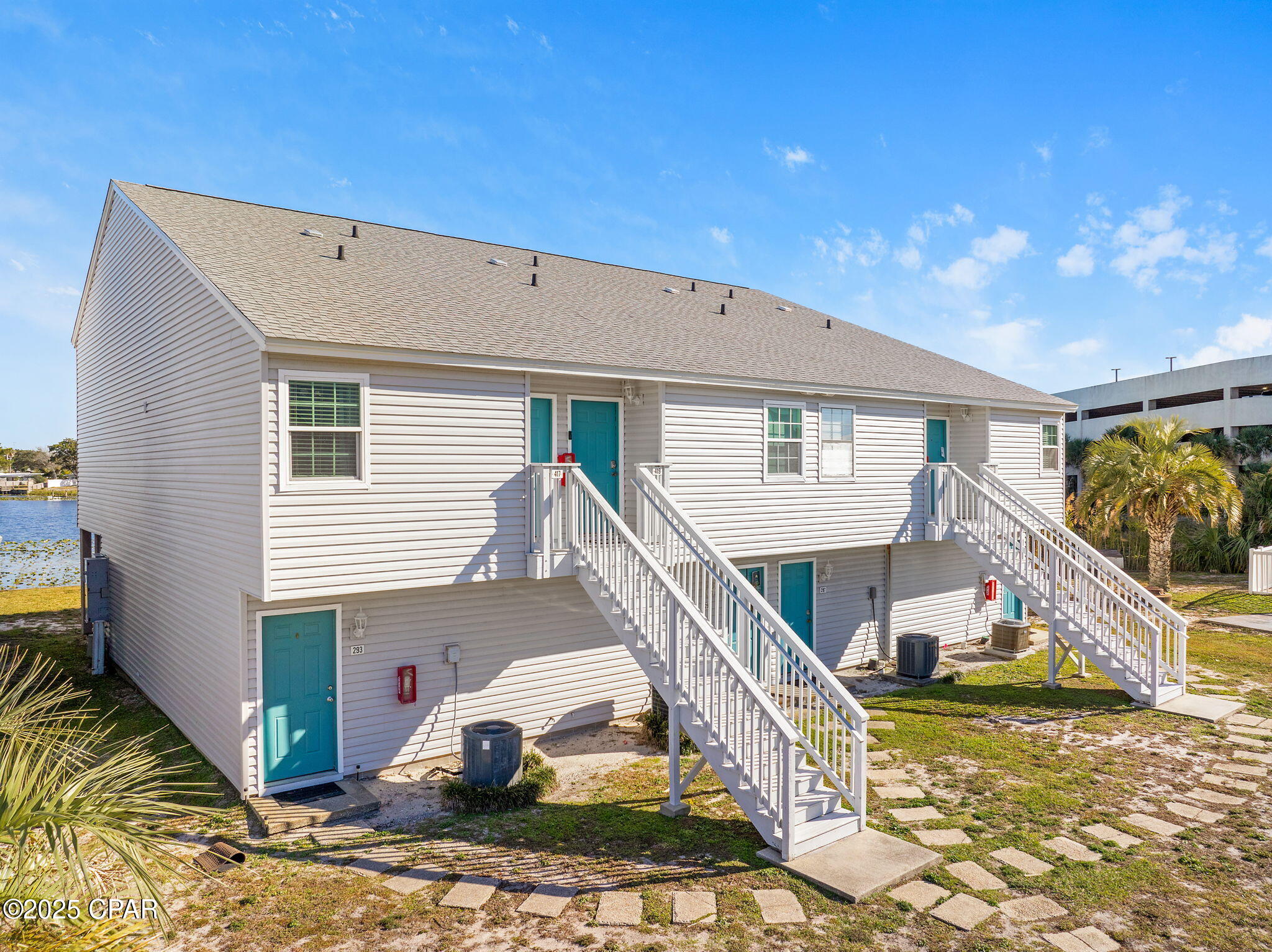 Two-story waterfront building with teal doors, white exterior stairs, and palm trees.