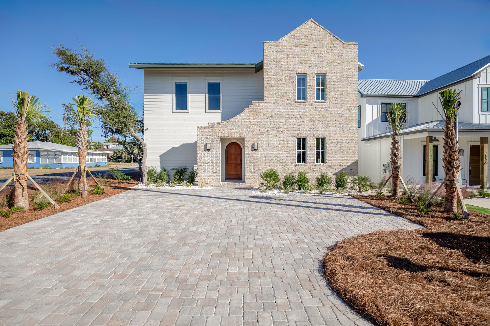 Two-story home with a pale brick facade, palm trees, and a broad paved driveway.